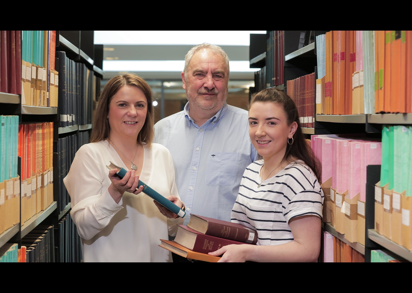 Image shows a woman and man, with a younger female student in a library. The younger woman is holding a number of different books in her hand.