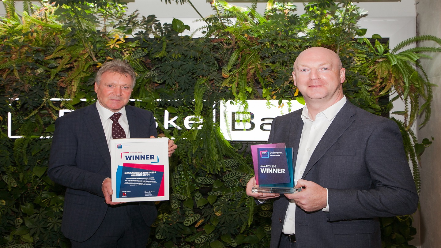 Two people standing in front of a Danske Bank green living wall, made up of plants and a lightbox Danske Bank logo. One person is holding a framed certificate and the other is holding an award.