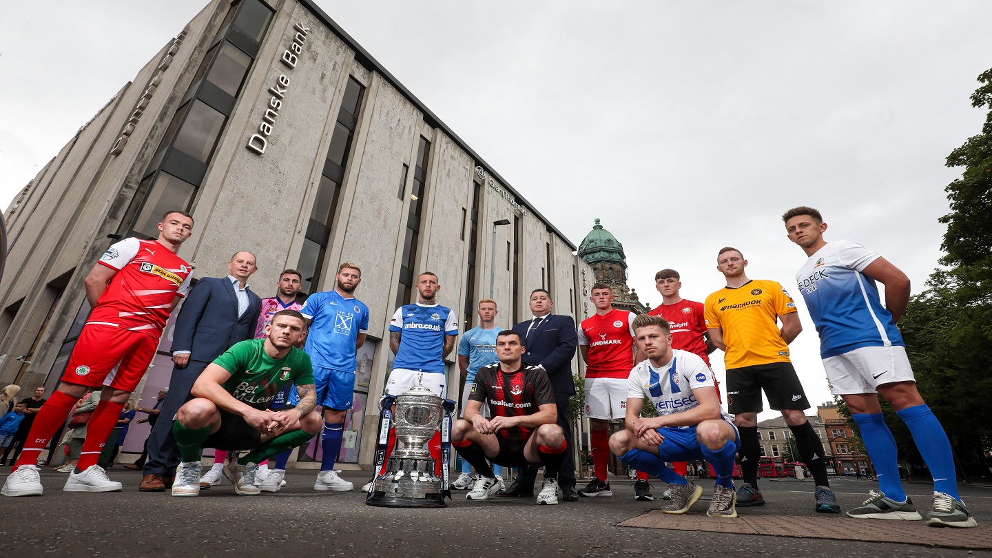 Fourteen people standing outside in front of Danske Bank headquarters. The Danske Bank Premiership trophy is sitting in front of them. Tweleve are players in full kits.