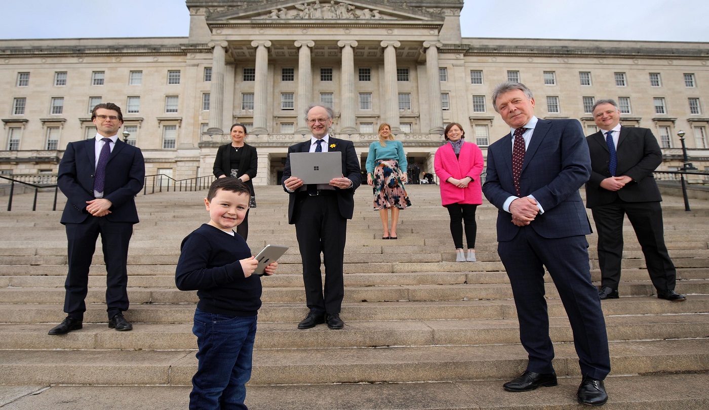 Seven adults and one young boy stand outside, on the steps in front of Stormont Parliament buidlings. The young man is holding a tablet, and the man in the middle of the group is holding a laptop. All are smiling and looking down at the camera.