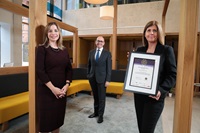 Three people stand, distanced from each other, inside. They are all wearing business-smart attire, and smiling. One woman to the front is holding a framed certificate.