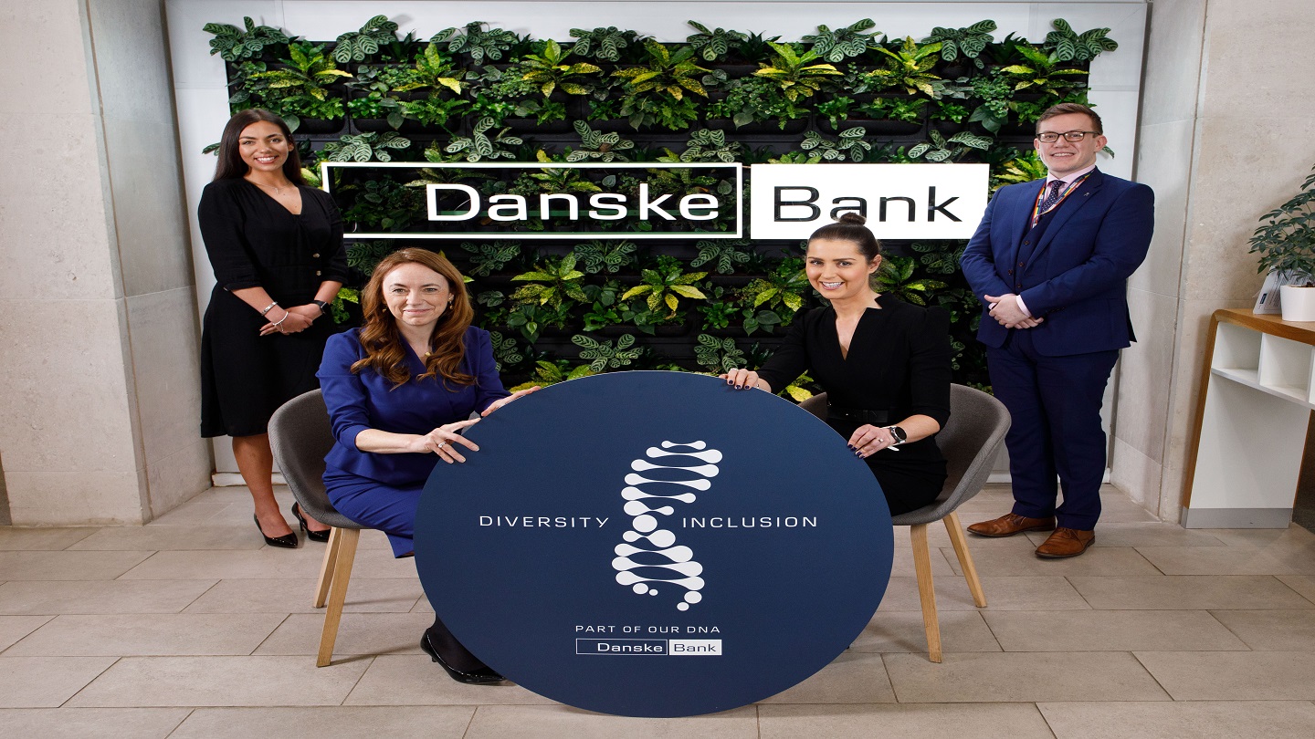 Four people inside a Danske Bank branch. Two at the front are siting and holding a circle sign with says 'Diversity and Inclusion, Part of our DNA'.