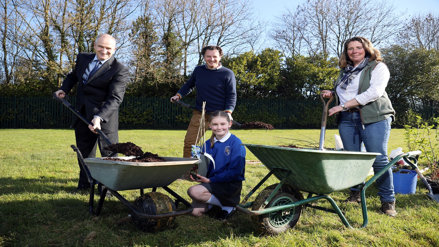 Three adults and a child in a school uniform outside doing some gardening with spades, wheelbarrows and plants.