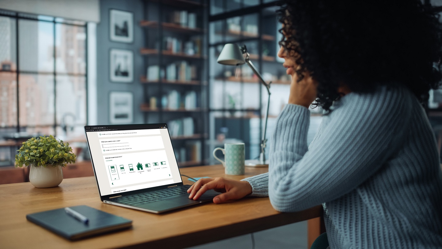 A person sits at a table with a laptop open in front of them. On the screen is the Danske Bank Energy Trust Savings tool.
