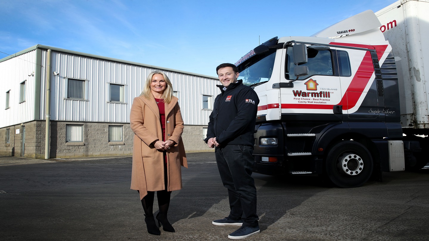 Two people stand outside, with a large lorry and building behind them.