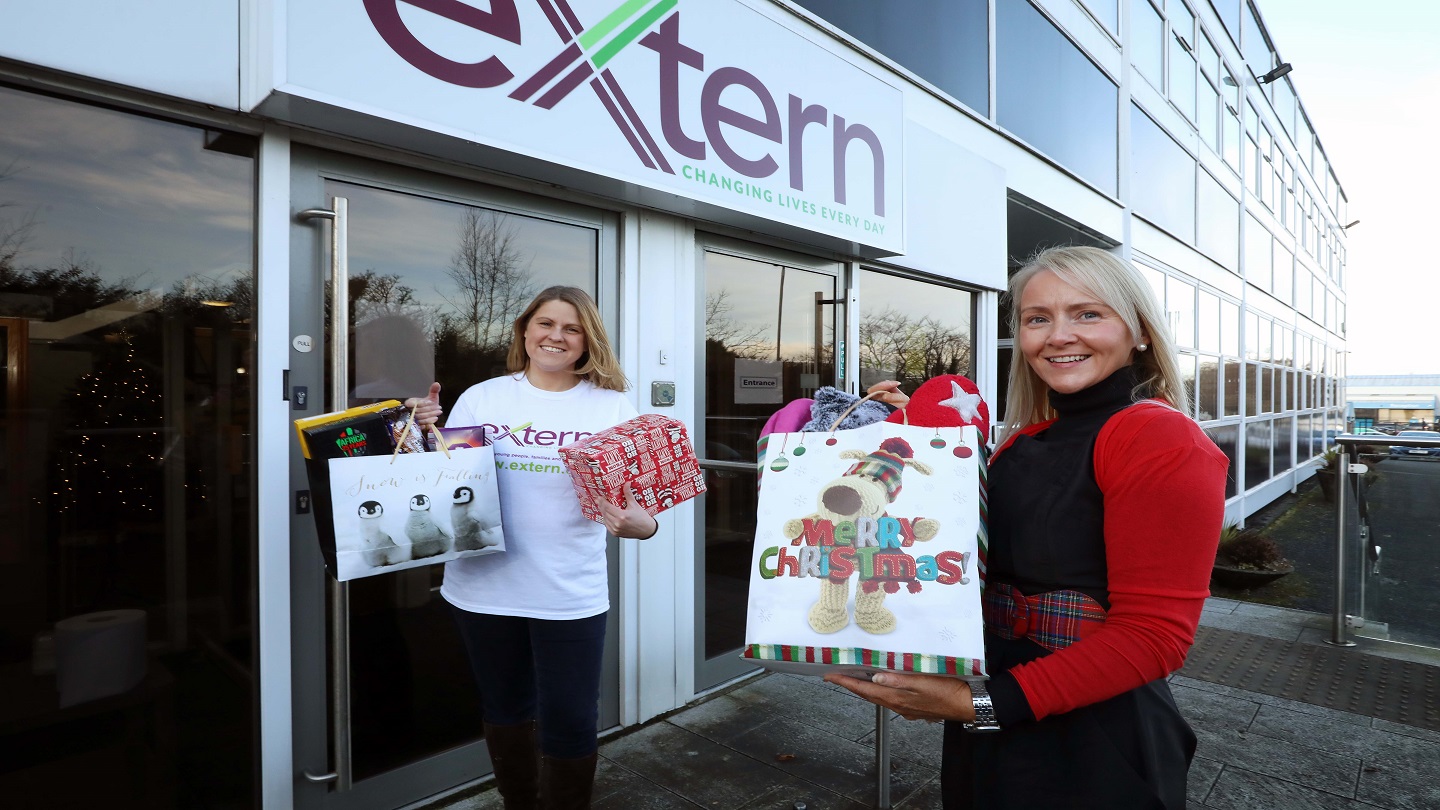 Two women standing outside a building with a white sign reading 'Extern'. They are both holding full Christmas gift bags and presents.