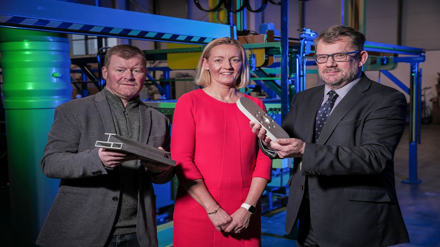 Two men and a women in a factory, the two men are holding some metal pieces of equipment.