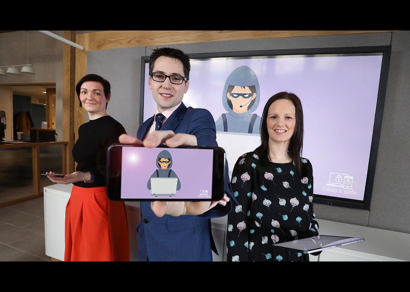 Two women stand either side of a man. They are holding tablets. The man is holding a phone to the camera, and on the screen is a purple background with a suspicious character on a computer. The same image is on a large TV behind the people.