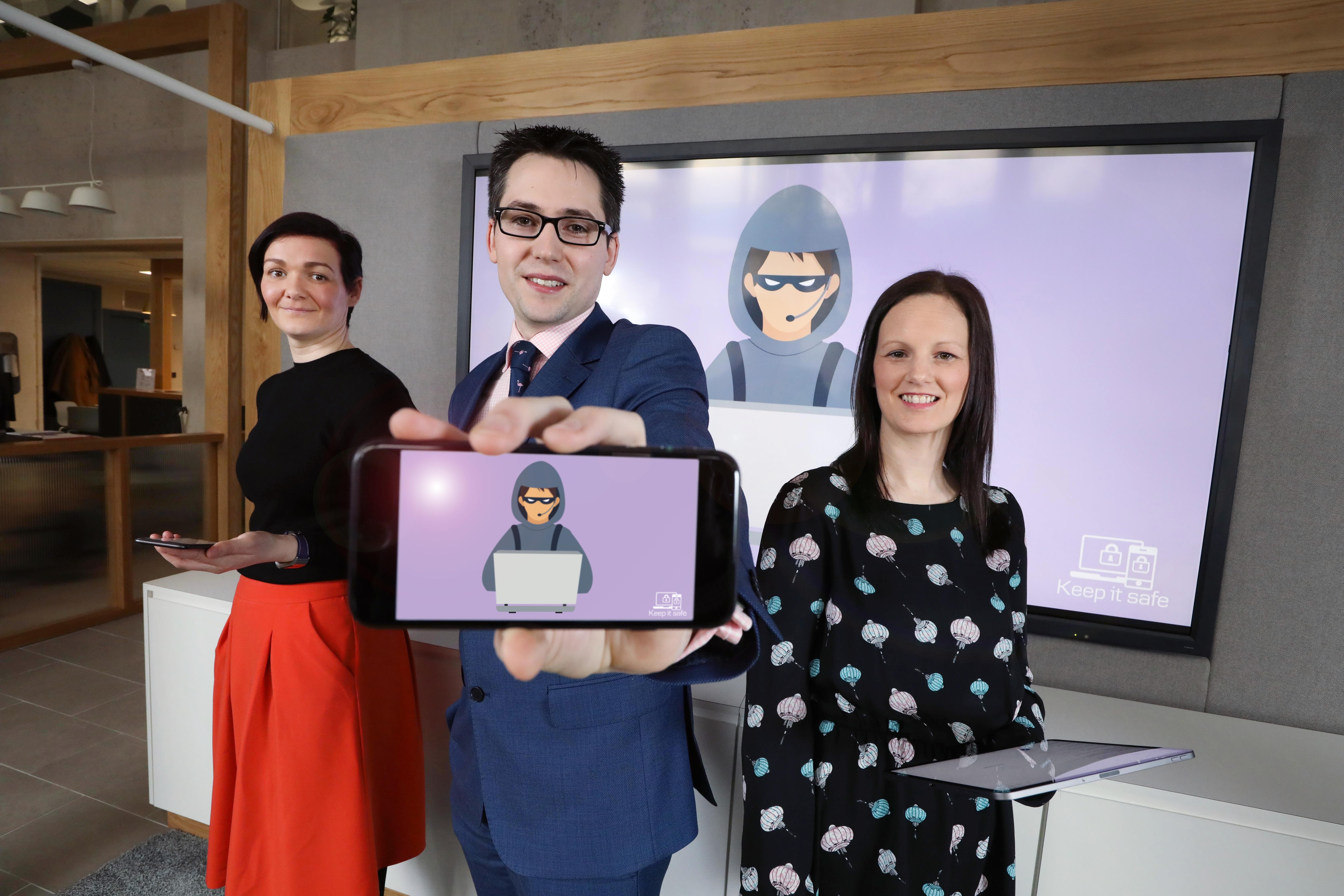 Two women stand either side of a man. They are holding tablets. The man is holding a phone to the camera, and on the screen is a purple background with a suspicious character on a computer. The same image is on a large TV behind the people.