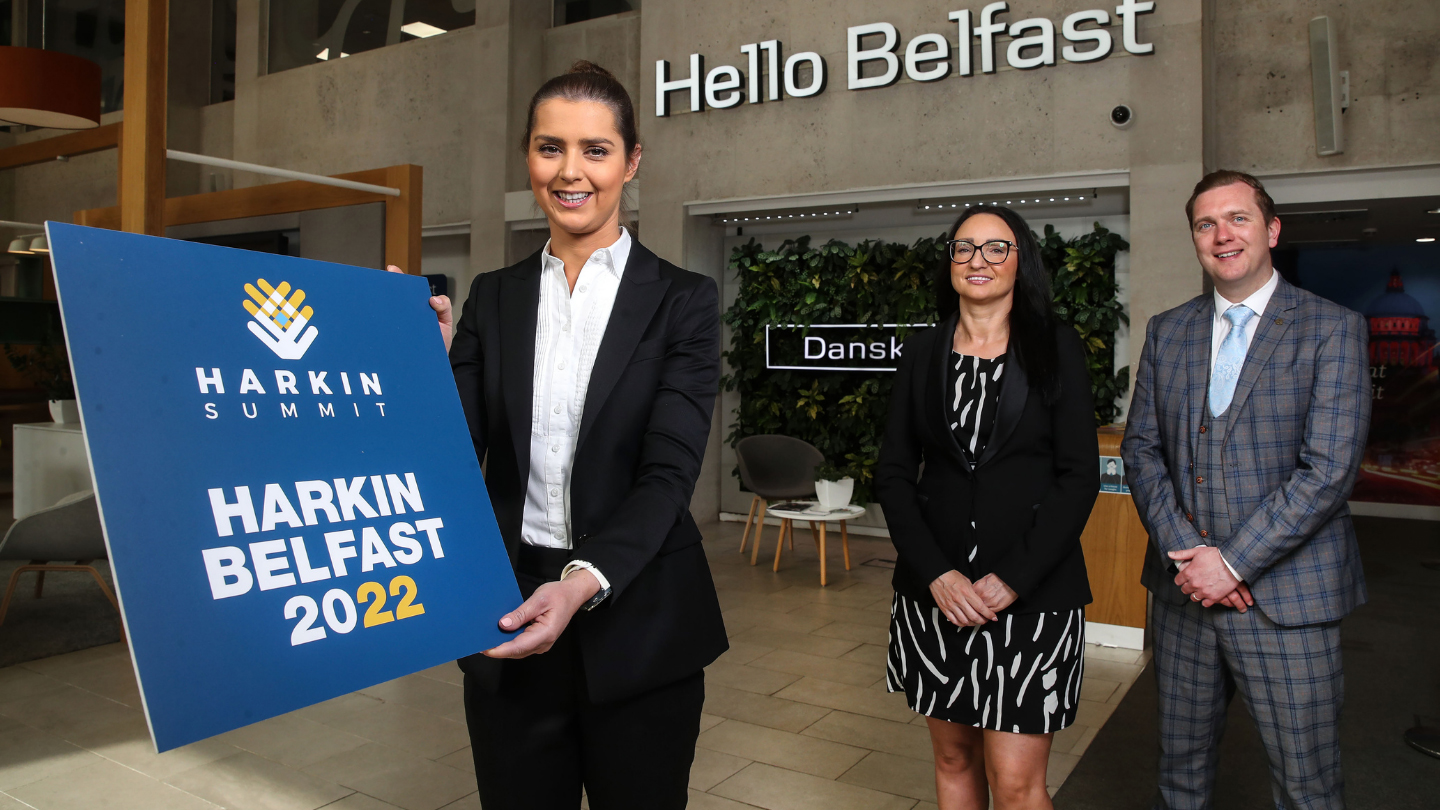 Three people standing inside a Danske Bank branch. One at the front is holding a navy sign that says 'Harkin Summit Belfast 2022'.