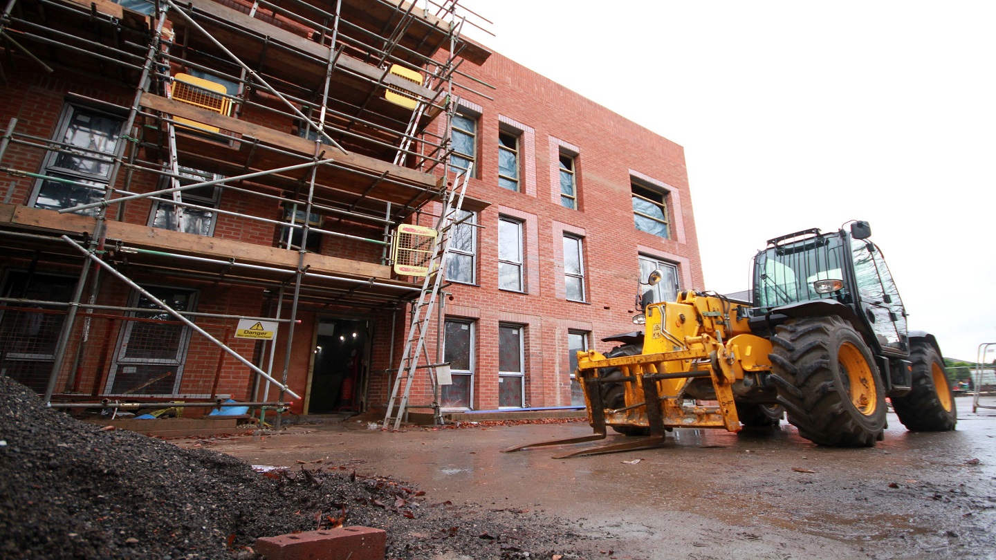 A yellow tractor sits outside a housing development building site. Half of the red bricked building with windows is up, and the other half is behind scaffolding.
