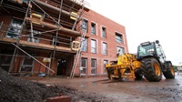 A yellow tractor sits outside a housing development building site. Half of the red bricked building with windows is up, and the other half is behind scaffolding.