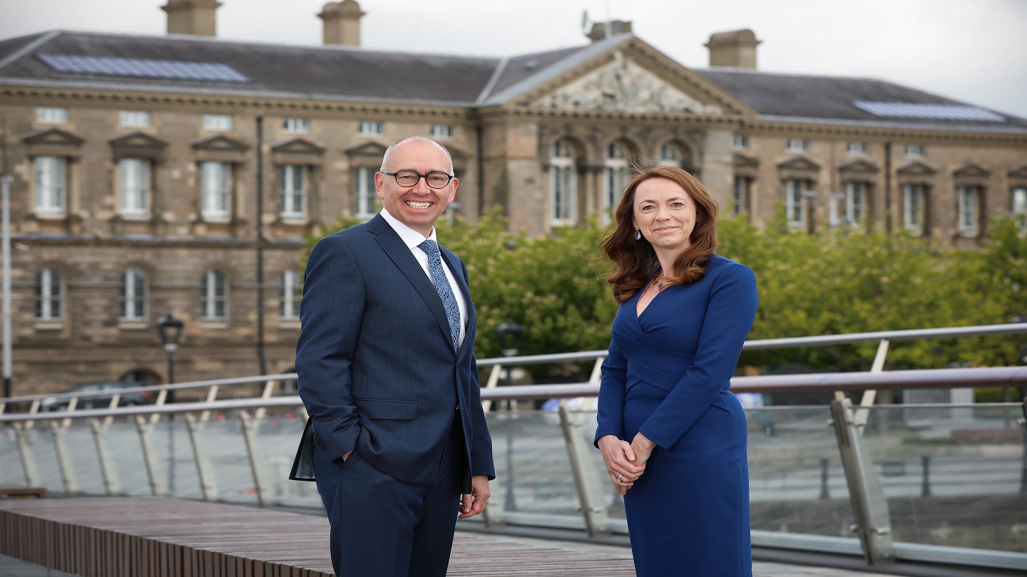 A man and woman in business clothes stand outside, both are smiling and looking at the camera.