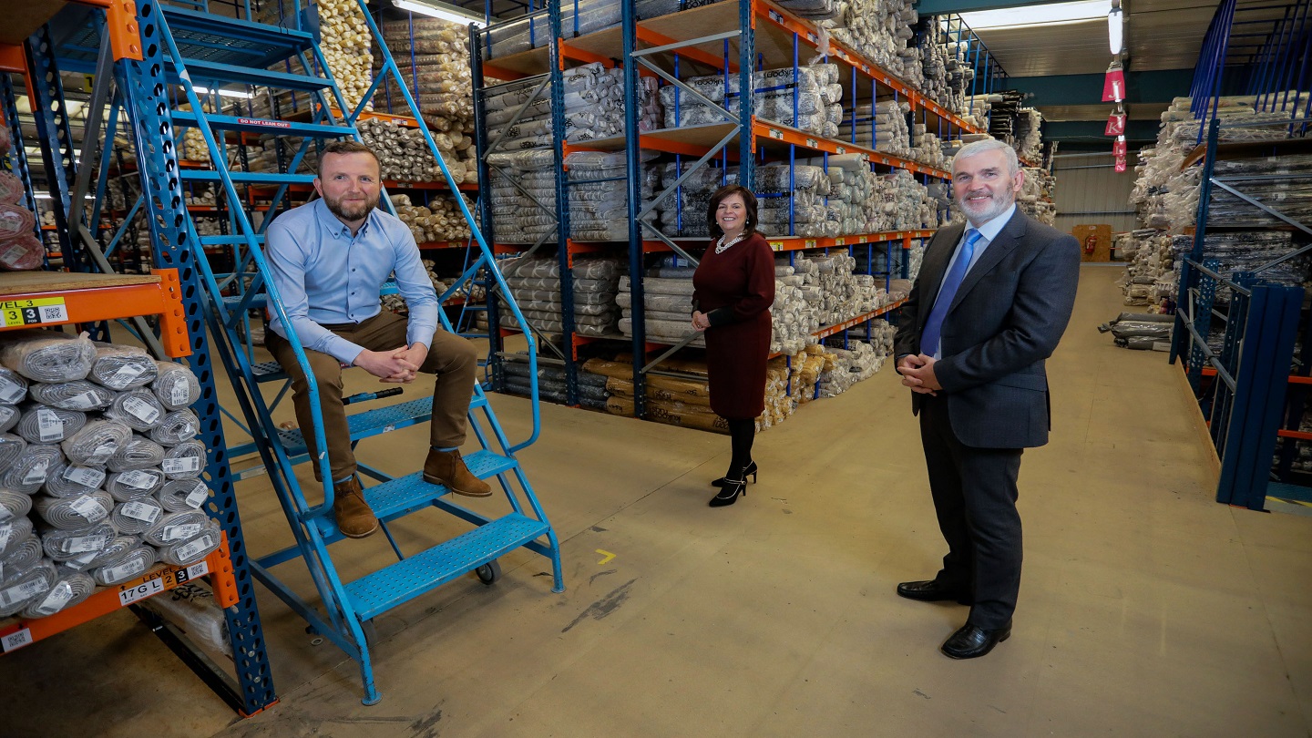 Three people inside a large warehouse, surrounded by rugs and carpets.