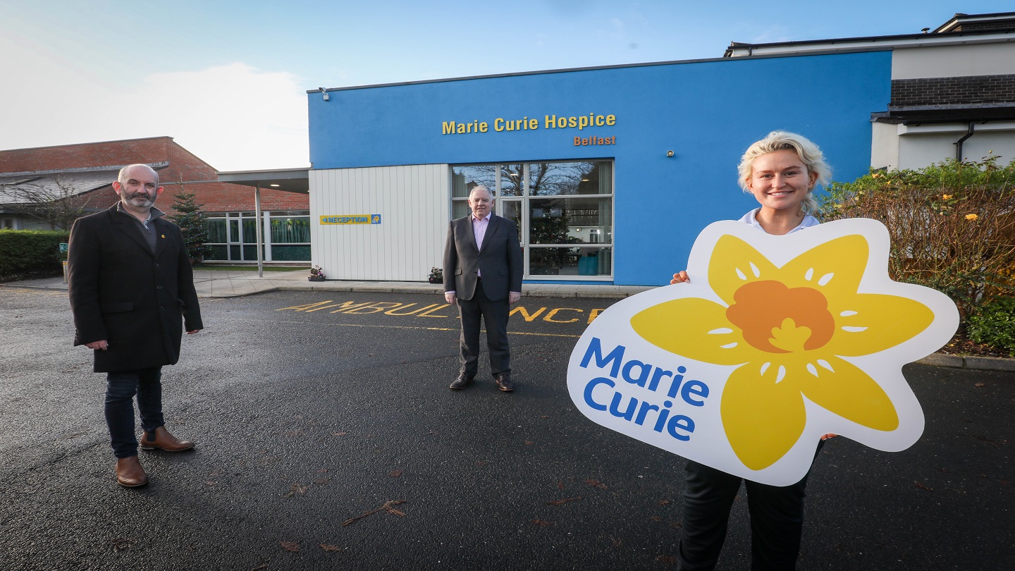 Three people standing outside the big blue 'Marie Curie Hospice' building in Belfast. There are two men in business wear and a nurse stands up front, she's holding a Marie Curie sign with their signature yellow daffodil.