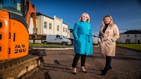 Two women standing outside on a housing development. A yellow piece of machinery is towards the front left of the frame.