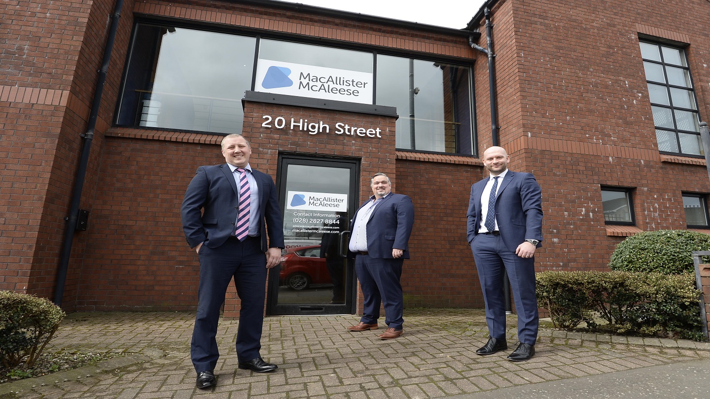Three men in suits stand outside a building with a sign 'MacAllister McAleese'.