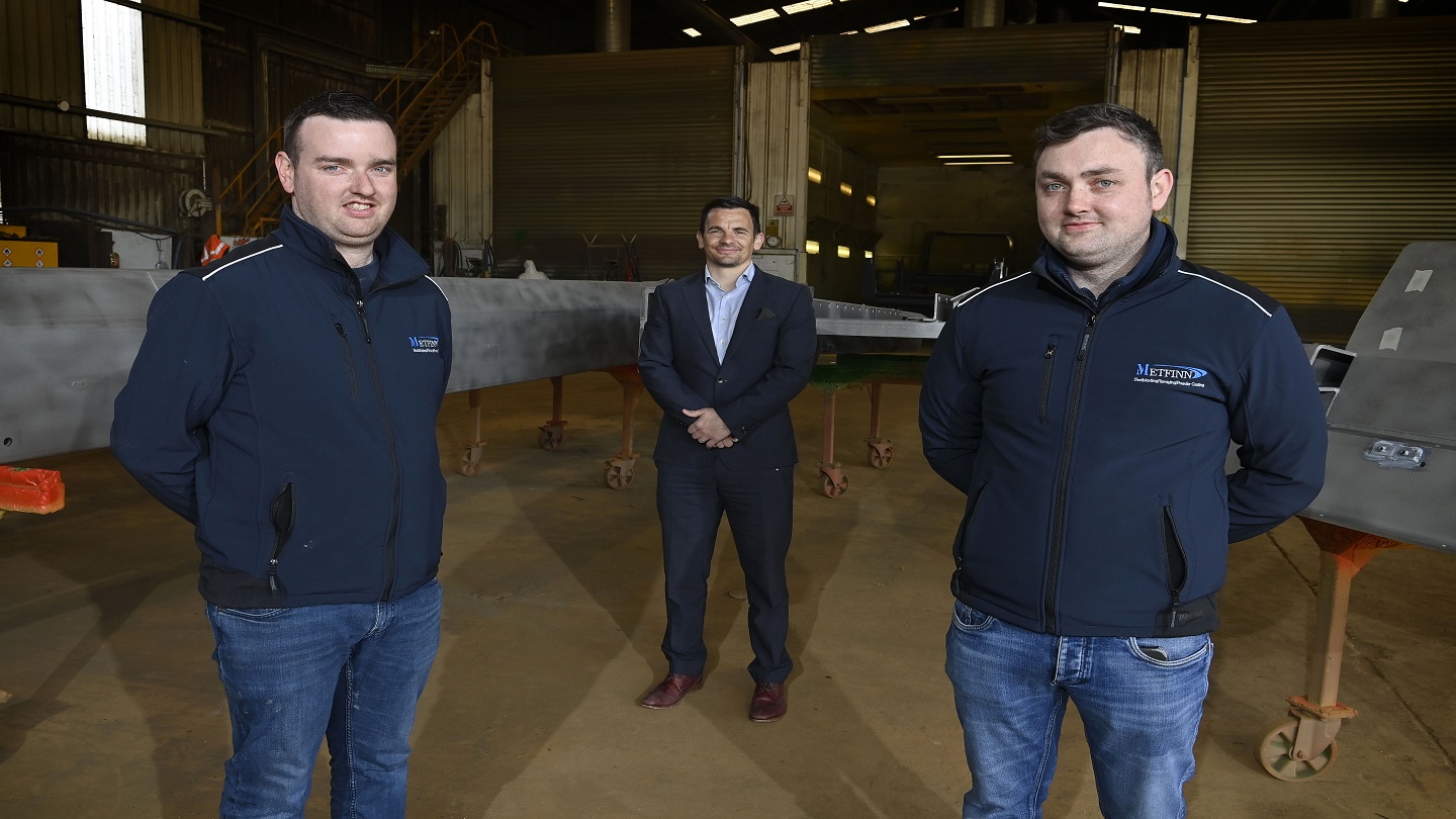 Three people standing inside a large factory, there's machinery behind them.