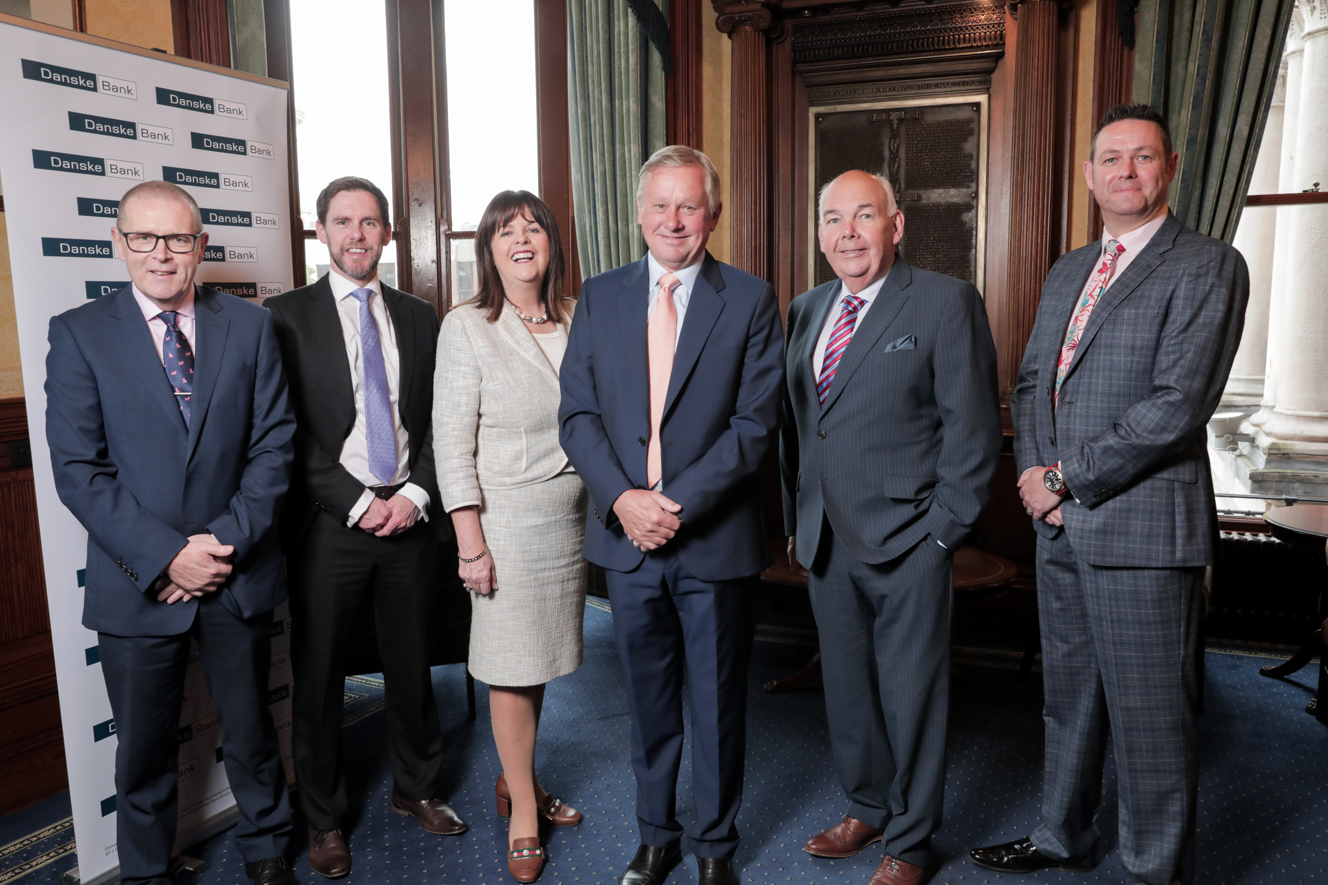 Six people, smartly dressed, stand beside each other looking into the camera and smiling. Behind them to the left is a Danske Bank branded pull up banner.