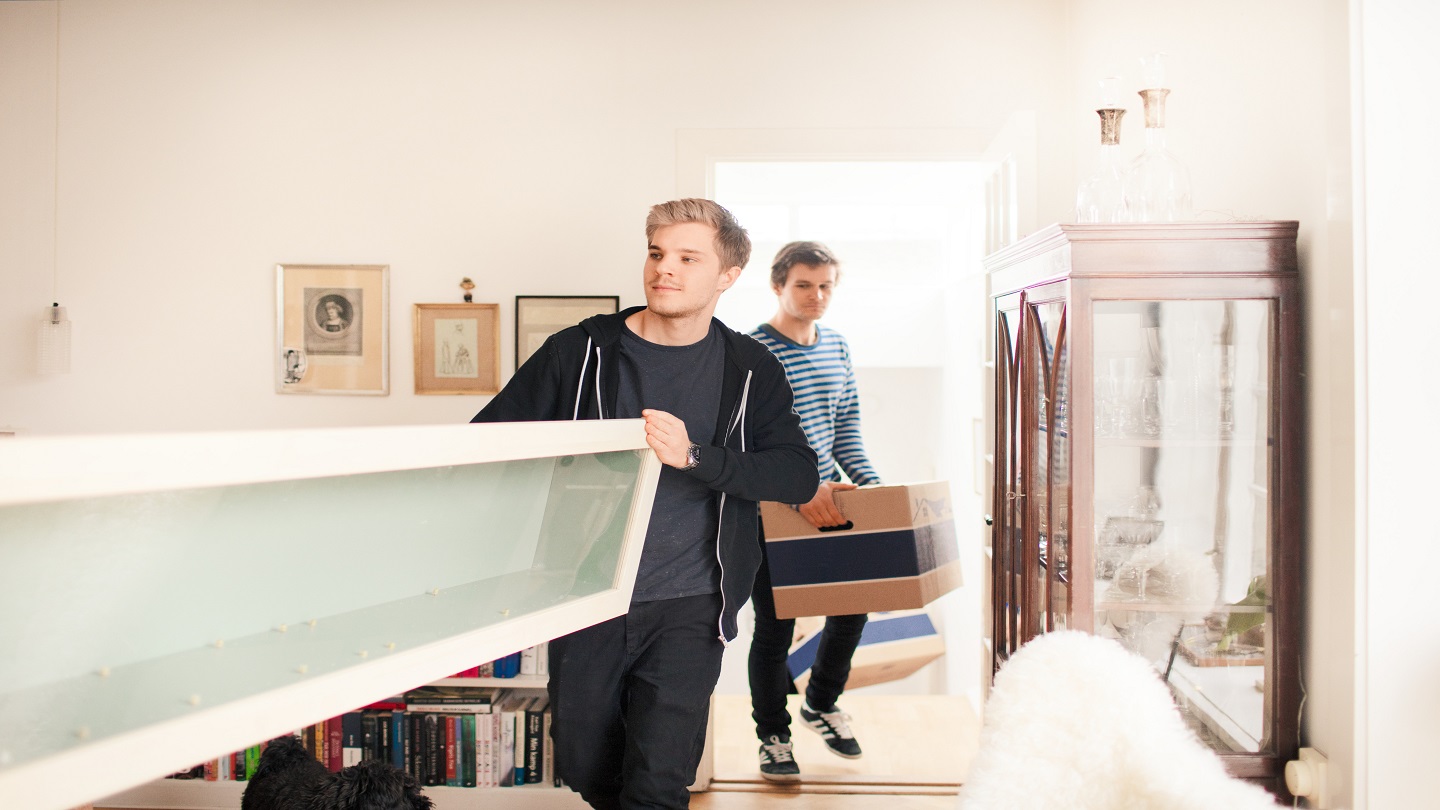 Two people moving into a house - one is carrying a large shelf with someone else off screen, and the other is following behind with a moving box.