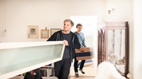 Two people moving into a house - one is carrying a large shelf with someone else off screen, and the other is following behind with a moving box.