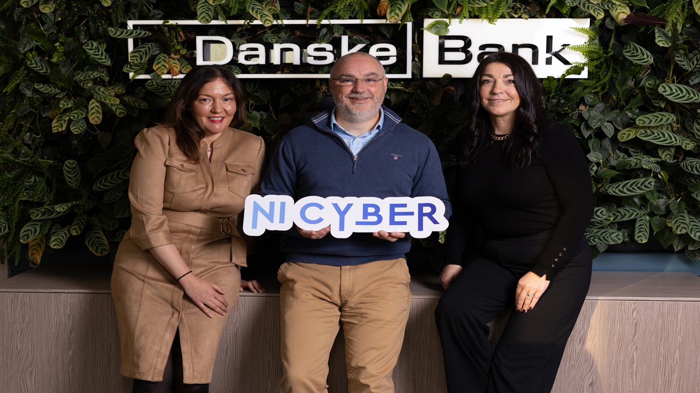 Three people in a Danske Bank branch in front of a Danske Bank sign, holding an NI Cyber logo.