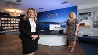 Two women stand inside an Oasis Travel agents. They stand socially distanced in the office, and there is a photo of a blue sky and beach at the reception desk behind them.