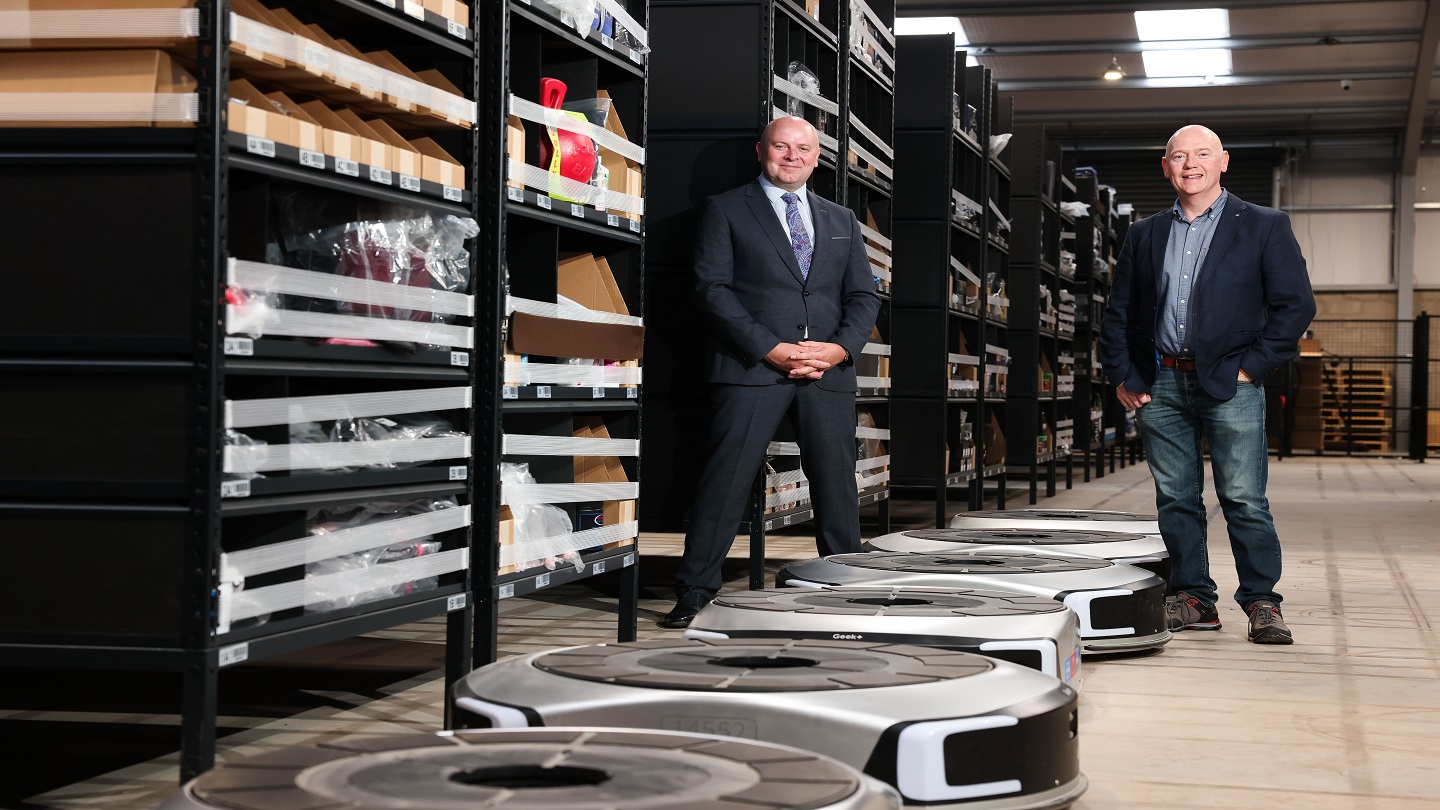 Two people standing inside a large storage warehuse. In front of them are a number of large, cicular robots.