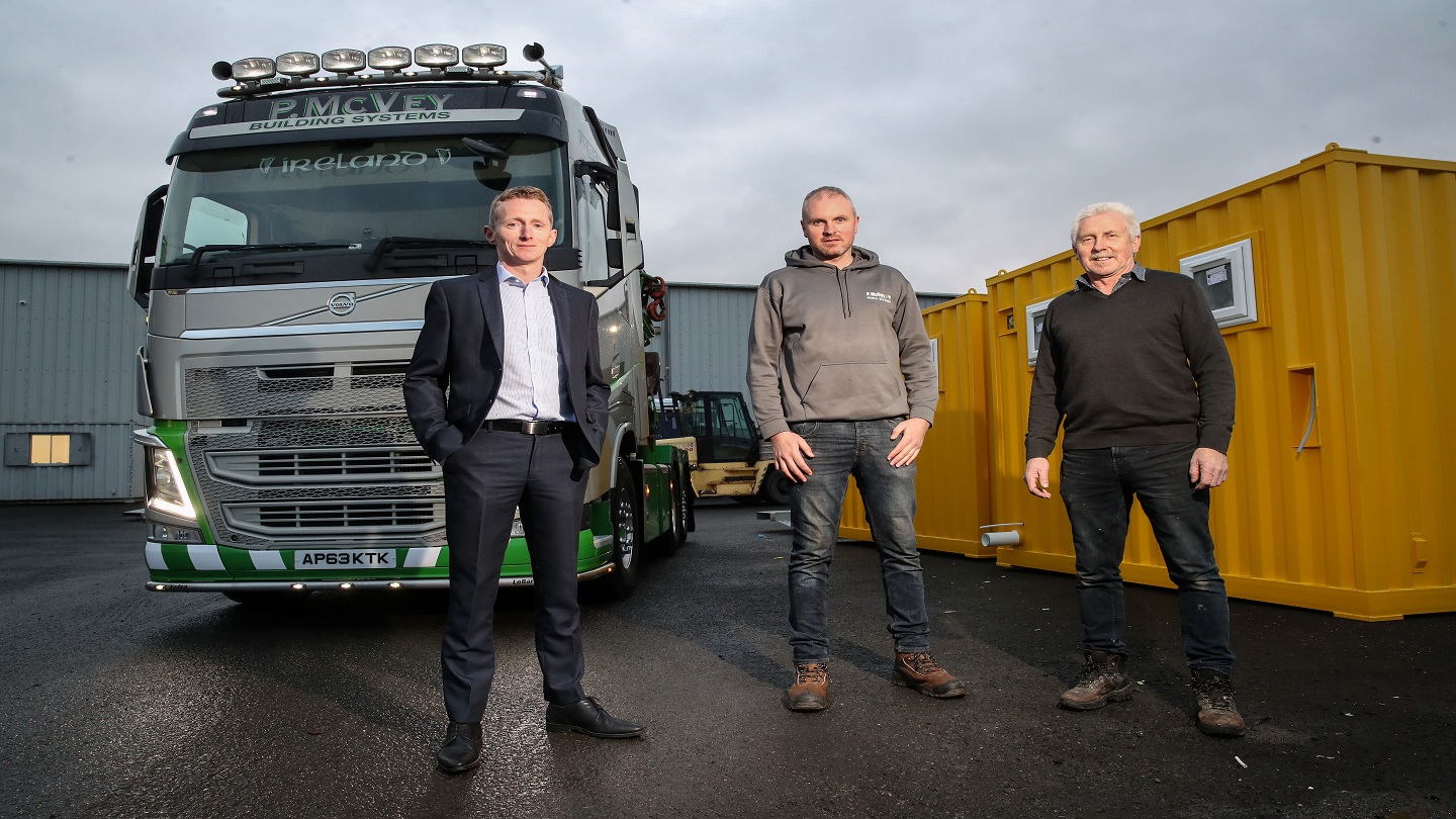 Three men stand outside. Behind them is a HGV and a mobile storage unit.