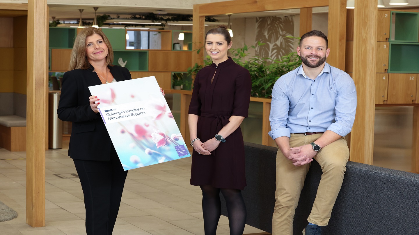 Three Danske Bank colleagues in a branch looking at the camera.  One colleague is holding a prop that reads 'Guiding principles on Menopause Support'.