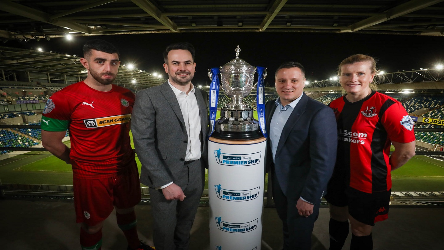 Four people stnding on a balcony in a sports pitch. Two are in full football kits (both red) and two are in suits. In the middle is a big silver trophy with blue and white Danske Bank ribbons sitting on a plinth that has the Danske Bank Premiership logo on it.