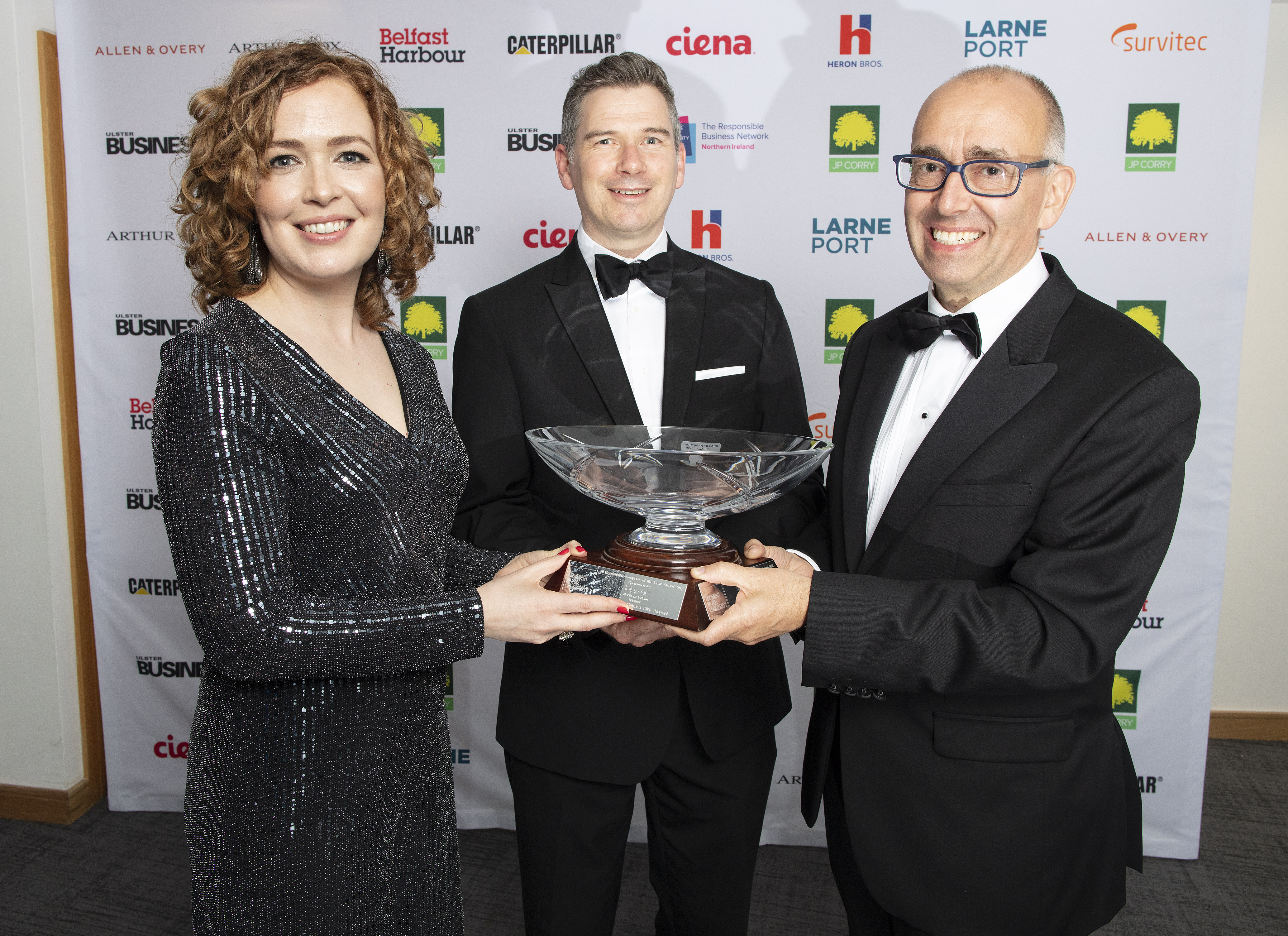 Three people stand at an awards ceremony, all very smartly dressed, they are holding an award - a big commemorative glass bowl.. There is a media backdrop with lots of logos behind them. 