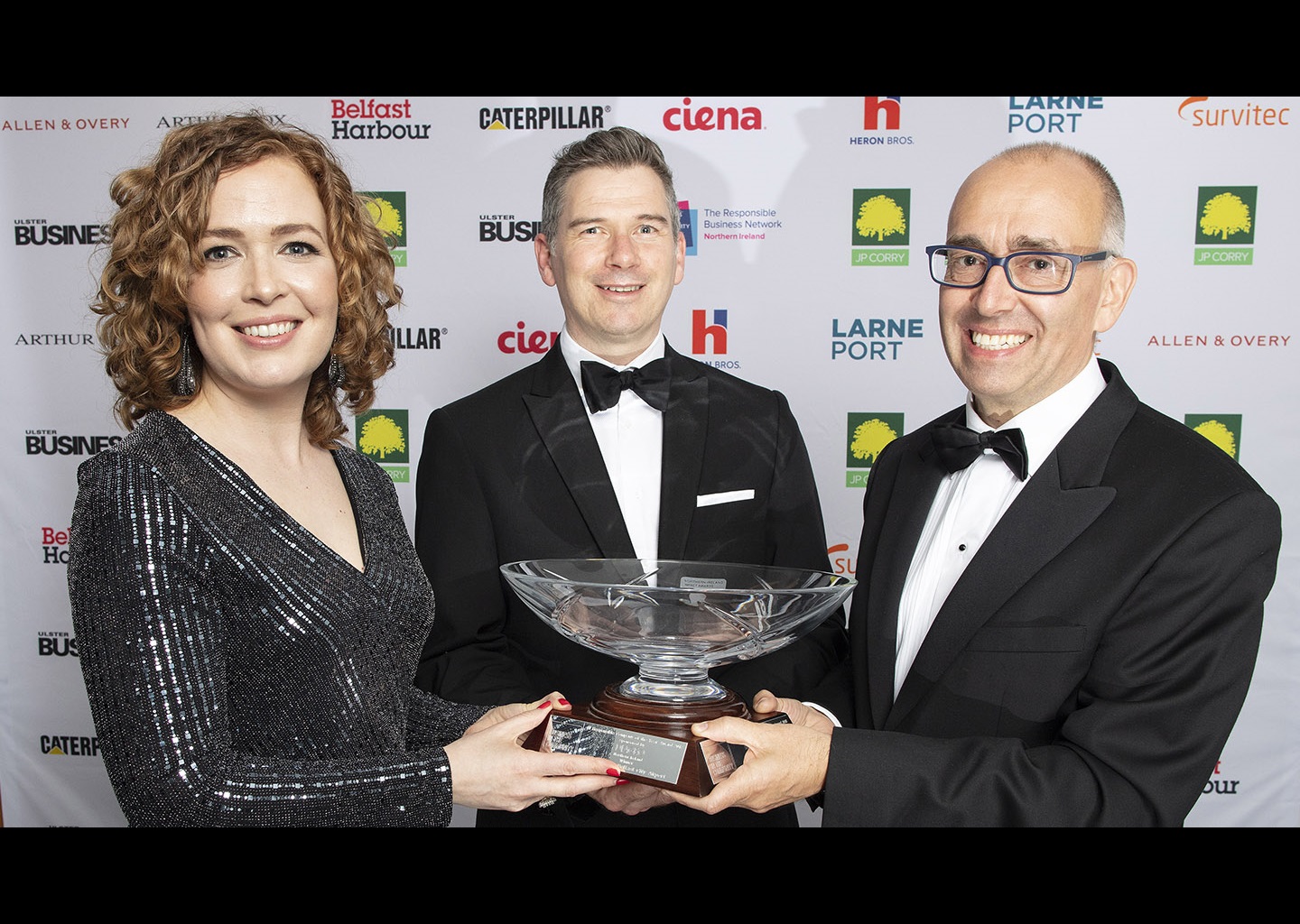 Three people stand at an awards ceremony, all very smartly dressed, they are holding an award - a big commemorative glass bowl.. There is a media backdrop with lots of logos behind them. 