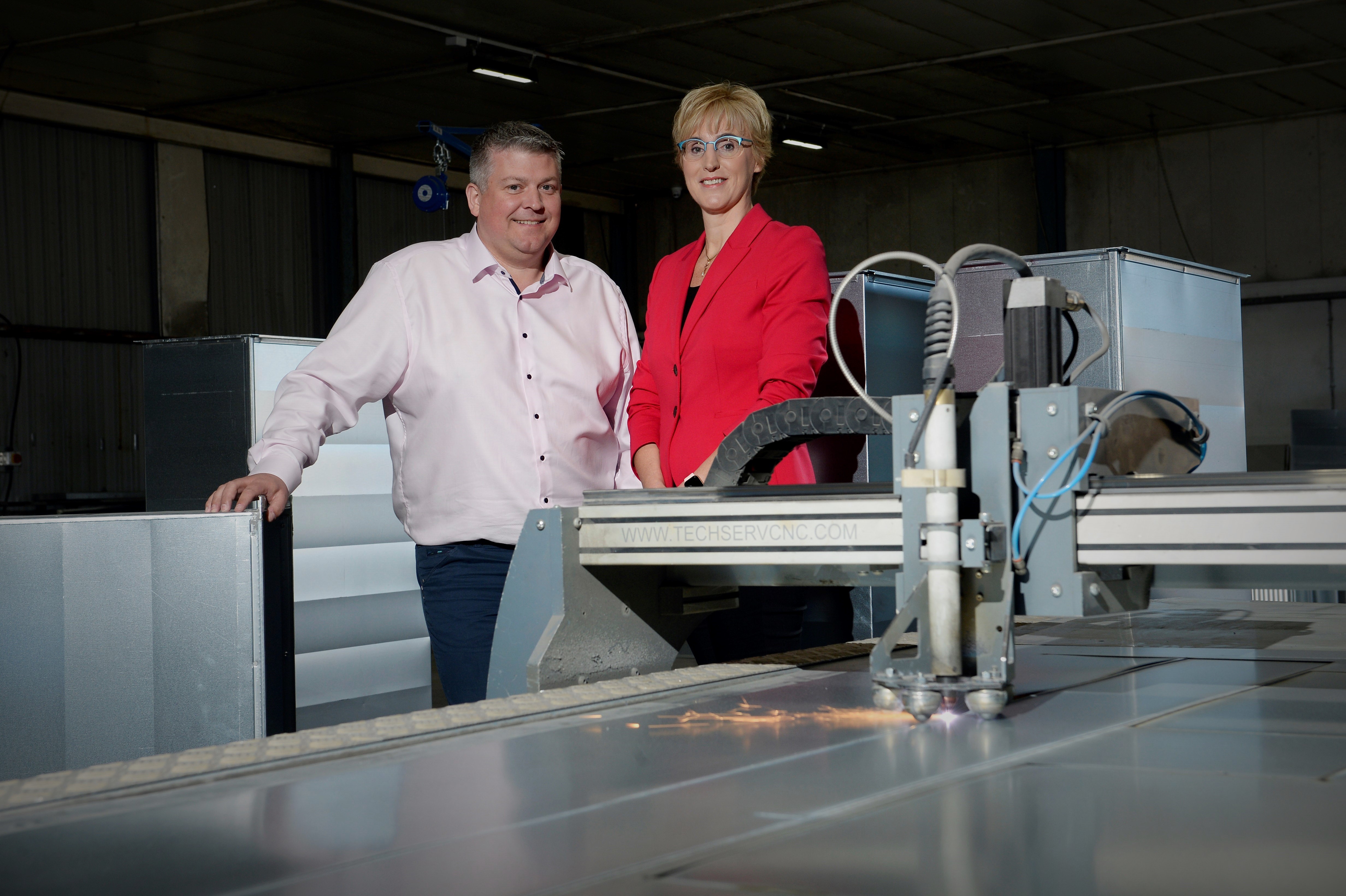 A man  and woman stand amongst some machinery, one of which is on an creating a little red spark.