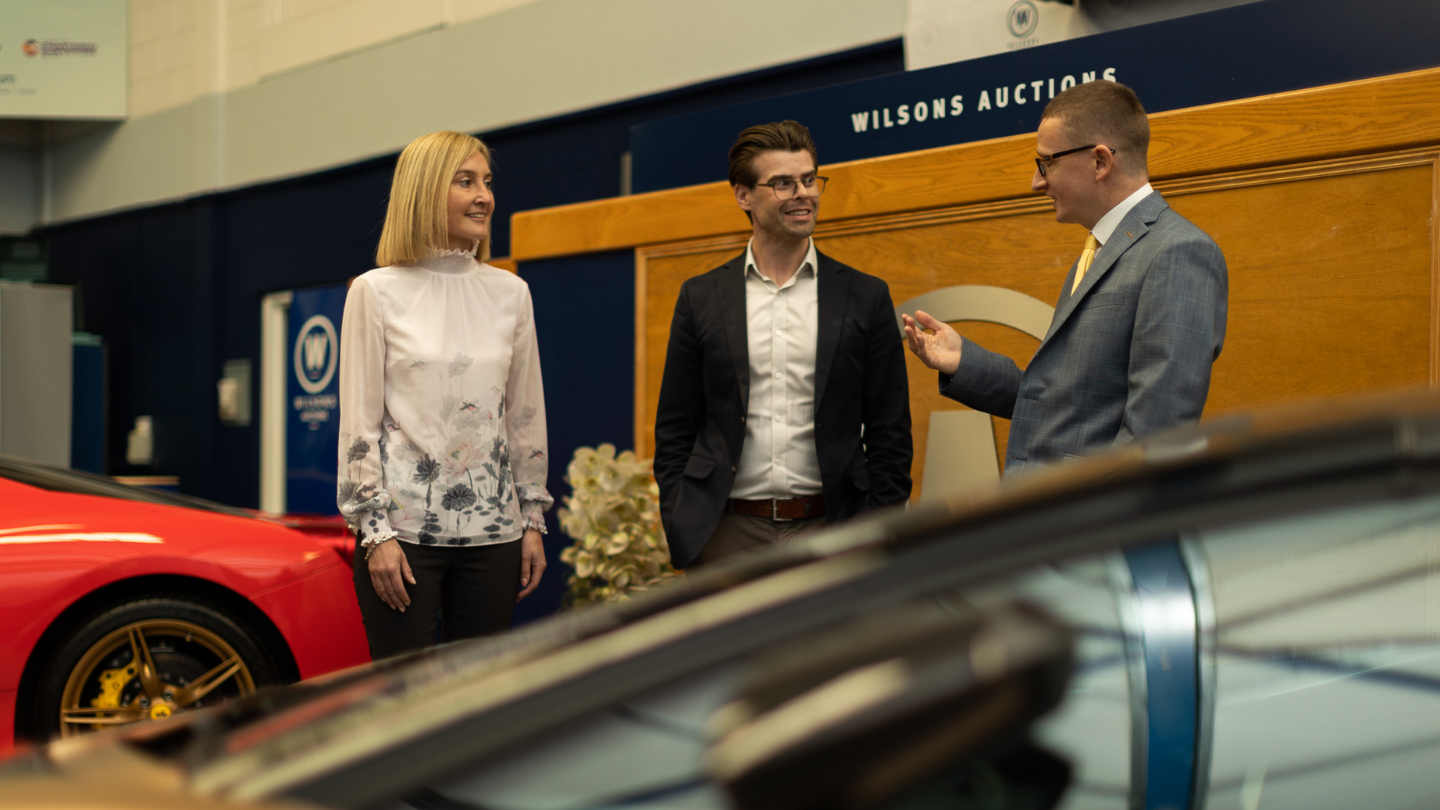 Three people stand inside a Wilsons Auction room. They are chatting, and around them are a couple of sports cars.