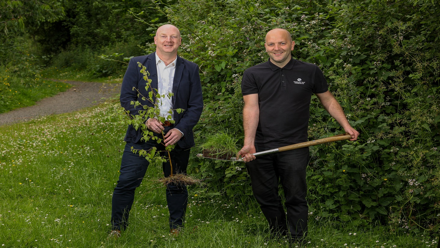 Two people stand outside in a green area. One is holding a tree sappling and the other has a spade with a lump of dirt on it, they are getting ready to plant the tree.