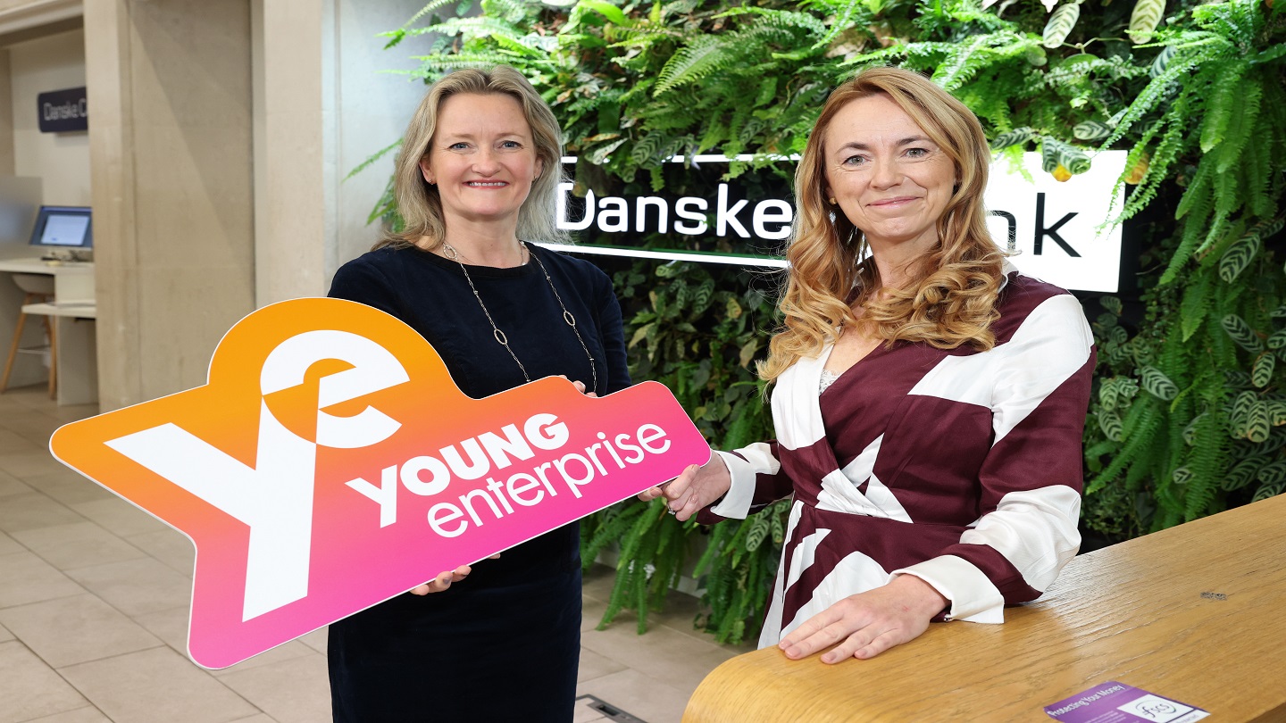 Two women stand in a Danske Bank branch, one is holding a Young Enterprise sign