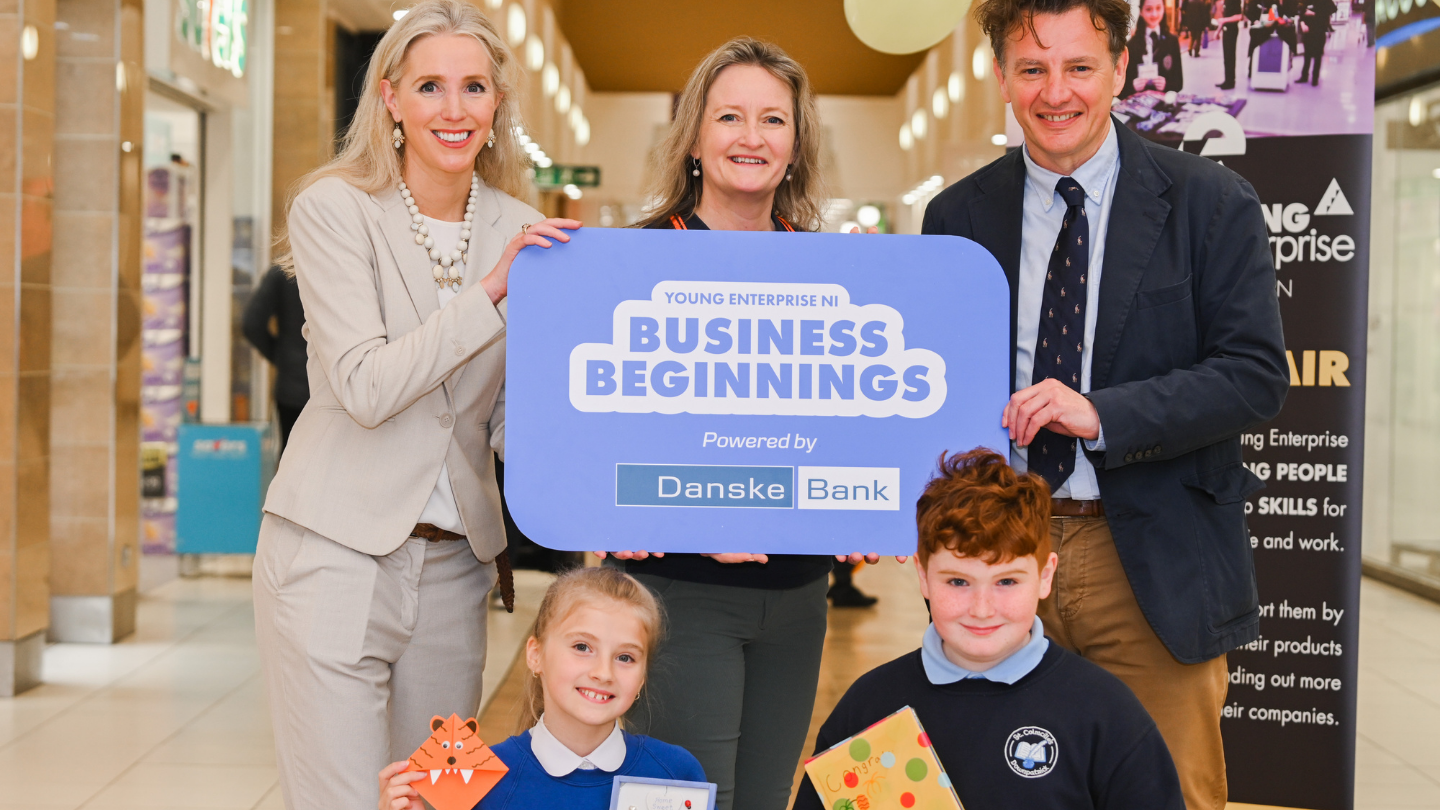Three adults and two school pupils standing in a shopping mall. They are holding a small sign that says 'Young Enterprise NI Business Beginnings, Powered by Danske Bank'.
