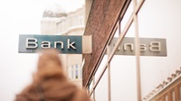 Image outdoors - out of focus is a person walking towards a bulding with a sign that says 'Bank'.